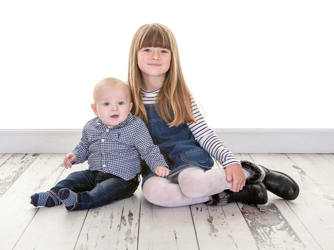 Sister wearing a stripey top and dungaree dress with her little baby brother wearing a checkered shit and jeans, sat on the floor together
